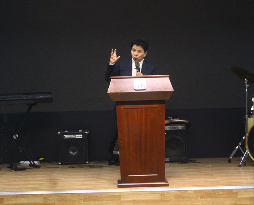 A person standing at a podium with a microphone Description automatically generated