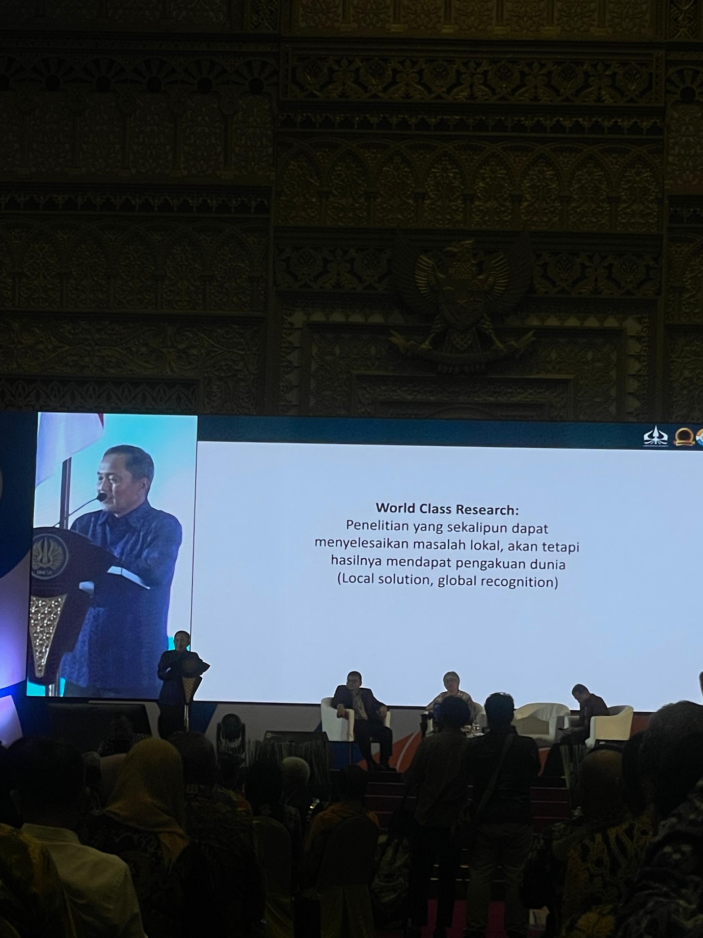 A person standing at a podium with a large screen Description automatically generated