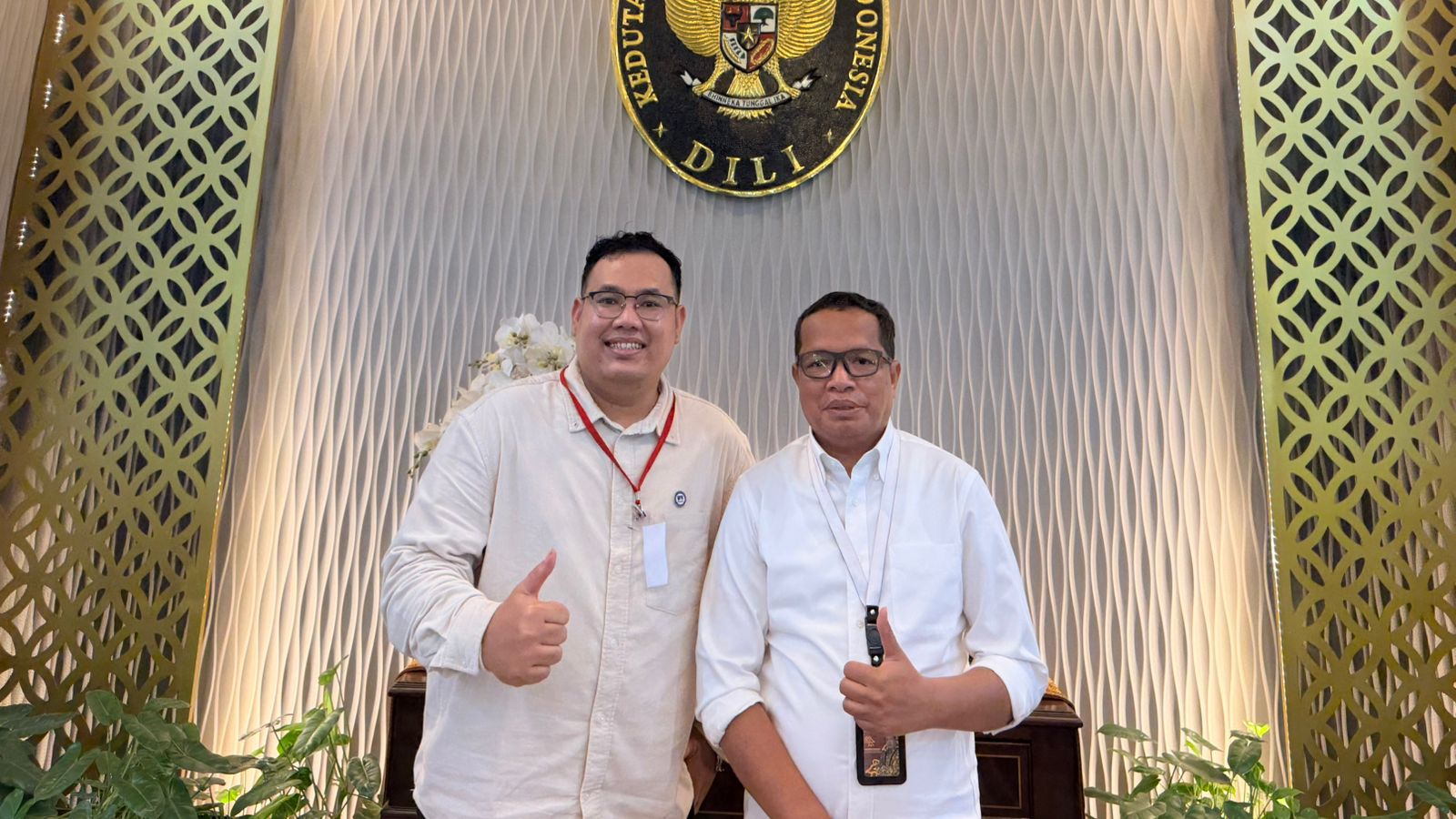 Two men standing in front of a wall with a sign Description automatically generated